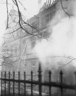 Fassade eines Gebäudes mit teilweise zerstörtem Dachstuhl. Vor dem Gebäude wabert Qualm.
