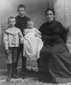 Photographie von Lina Sommer im mittleren Alter, sitzend in einem Stuhl, einen ihrer Söhne im Kleinkindalter in die Kamera halten. Daneben stehen zwei weitere Söhne im Kindesalter.