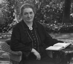 Photographie einer älteren Lina Sommer in einem Korbstuhl in ihrem Garten sitzend. Vor sich ein Tisch, auf dem ein aufgeschlagenes Buch liegt.