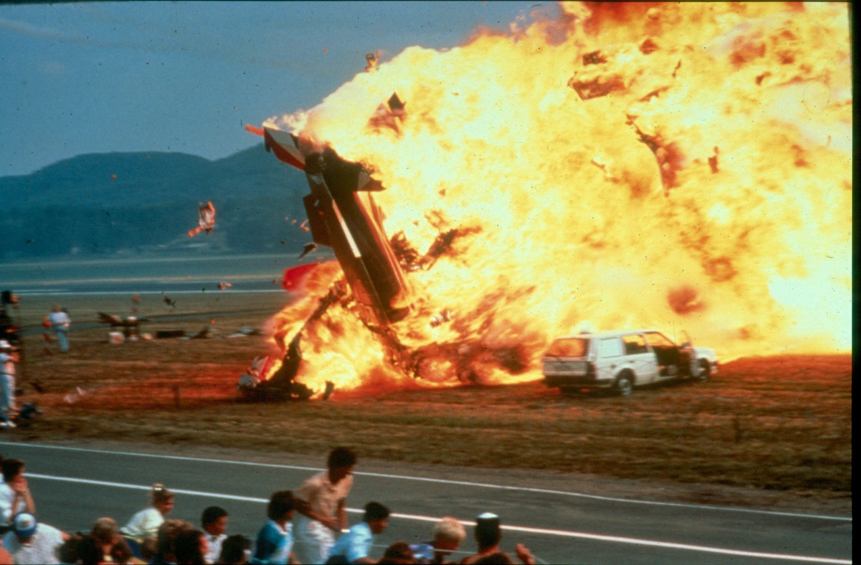 Man sieht einen auf ein Feld stürzenden explodierenden Jet und davor fliehende Menschen.